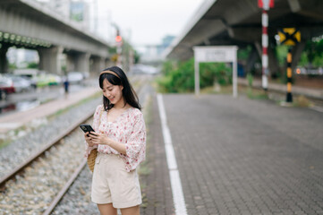 Fototapeta premium Asian young woman traveler with weaving basket using a mobile phone beside railway train station in Bangkok. Journey trip lifestyle, world travel explorer or Asia summer tourism concept.