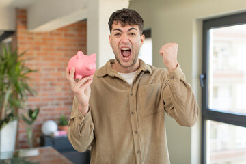 young handsome man feeling shocked,laughing and celebrating success. piggy bank concept