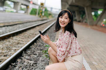 Asian young woman traveler with weaving basket using a mobile phone beside railway train station in Bangkok. Journey trip lifestyle, world travel explorer or Asia summer tourism concept.