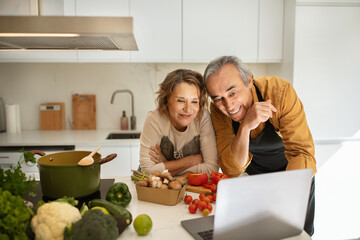 Happy senior spouses watching video recipe on laptop while cooking lunch in kitchen interior together, free space