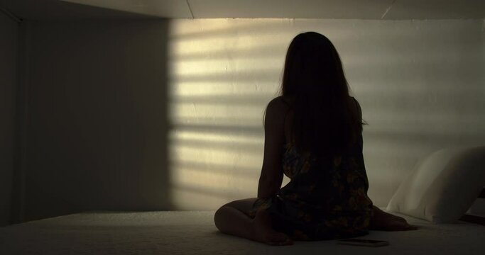A lonely woman is sitting on the bed in a narrow dark room. This concept use for symptoms of stress, major depressive disorder (MDD), psychotic symptoms.