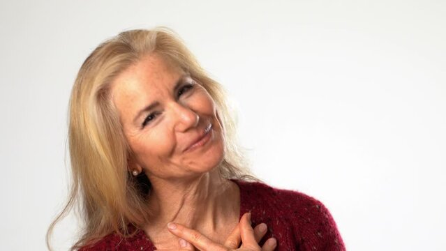 Closeup of middle age mature woman, receives beautiful compliments, finally raises her hands puts hands on chest. Beautiful mature blond isolated on solid white background. Copy space