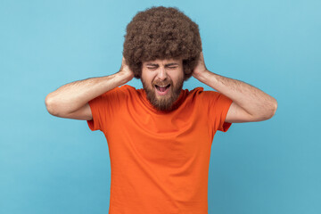 Portrait of man with Afro hairstyle covering ears, cannot focus in crowded place, feels discomfort over noise, being troubled to listen advice. Indoor studio shot isolated on blue background. - obrazy, fototapety, plakaty