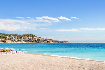 Fototapeta na wymiar Beach, sea and coast in Nice, South of France. French Riviera shore. Blue sky and turquoise water. Mountain village and town in the background. Bright summer landscape of mediterranean destination.