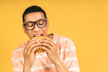 Hungry young african american man eating hamburger or burger isolated over yellow background. Diet...
