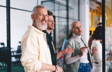 Half length portrait of cheerful bearded male entrepreneur smiling at camera during collaboration working process with blurred colleagues, successful Caucasian businessman posing in office interior