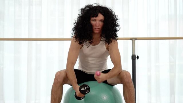 Guy with stage wig exercises with barbells sitting on ball