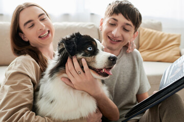 smiling and young gay men with tattoo cuddling Australian shepherd dog and holding photo album...