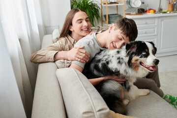 Cheerful same sex couple in casual clothes spending time and hugging furry Australian shepherd dog while relaxing on couch in living room at home