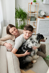 Young homosexual man hugging furry Australian shepherd dog near smiling long haired boyfriend while resting on couch in living room at home