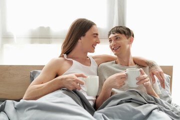 Smiling tattooed gay man in sleepwear hugging and looking at young happy boyfriend while holding...