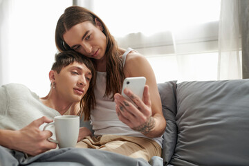 Young gay man in sleepwear holding coffee cup and looking at smartphone while sitting together with boyfriend on comfortable bed in morning time