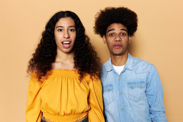 Young surprised couple standing against brown studio background with opened mouth and astonished facial expression, after hearing jaw dropping news or excited with sales and discounts