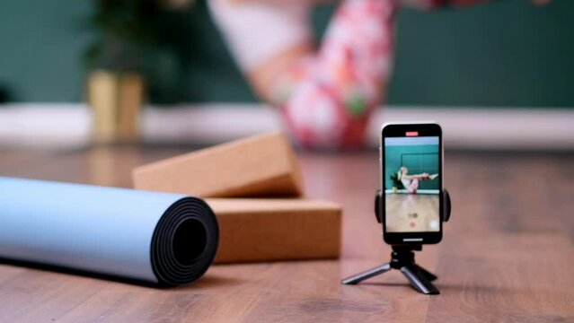 Professional yoga training in cozy studio tripod with phone and mat roll on floor against woman standing in pose female person recording exercise on camera to post on social media