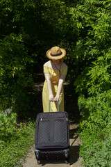 Asian girl in straw hat and yellow dress with big black suitcase among green trees. Summer vacation.
