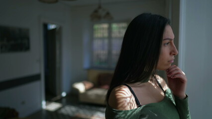 One contemplative young woman standing by window looking outside with pensive expression....