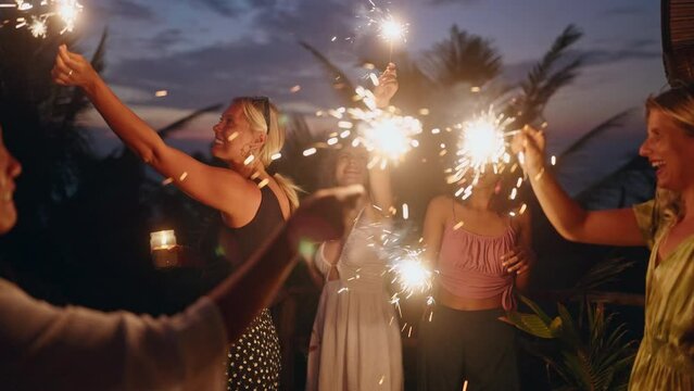 Young multiracial females have fun celebrating Christmas in tropics by sea with sparkling lights. Diverse happy women cheer and dance holding and waving sparklers at Independence Day party at seaside