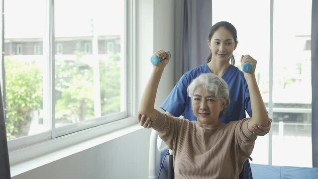 Young female physiotherapist talking and showing arm movements to gray-haired old woman using resistance bands for muscle exercise and physiotherapy