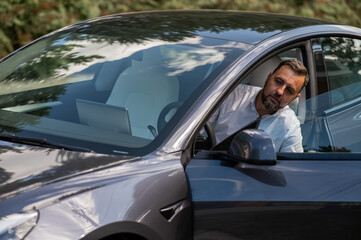 Caucasian bearded man in a suit gets out of a black electro car in the countryside in summer.