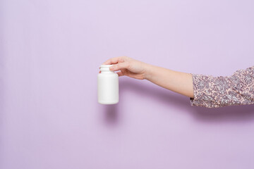 White plastic bottle in female hand in lilac background. Packaging for pills, capsules or supplements.