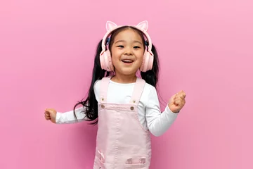 Foto auf Acrylglas Tanzschule little asian girl in pink children's headphones listens to music and dances on pink isolated background, korean child
