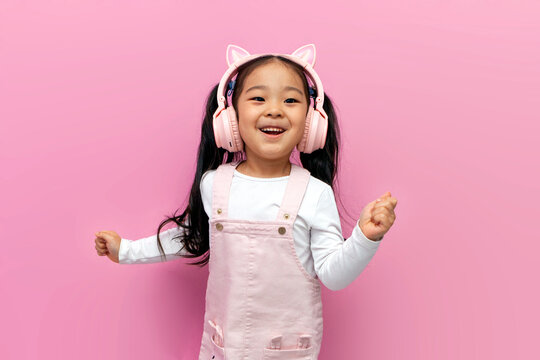 little asian girl in pink children's headphones listens to music and dances on pink isolated background, korean child