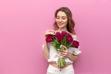 cute young woman in festive outfit holds bouquet of pink tulips and smells them on pink isolated background