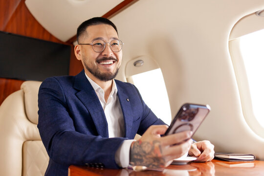 successful asian businessman in suit and glasses flies in private luxury jet and uses smartphone - Powered by Adobe