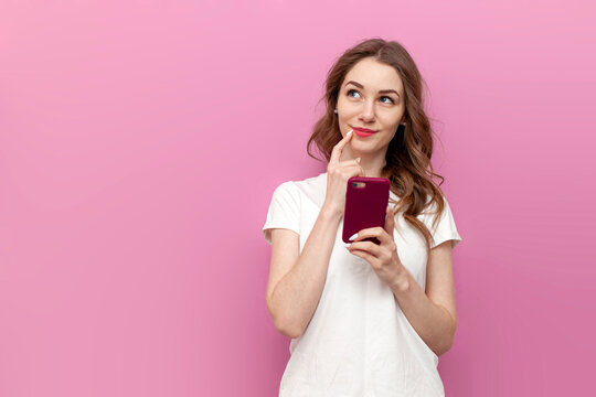 young pensive woman in white t-shirt uses smartphone and dreams on pink isolated background, girl uses the phone