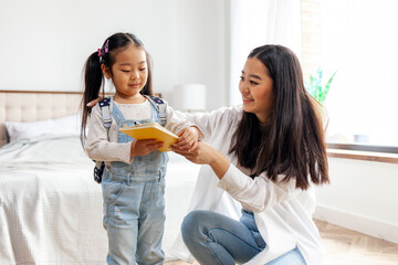 mother collects Asian daughter to school, woman puts on backpack with book to little girl, back to school