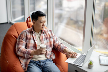 Motivated asian employee sitting on orange comfortable bean bag chair holding cup of aromatic coffee and working on laptop. Young male creating business project using modern wireless technology.