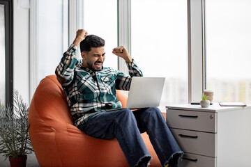 Excited indian worker celebrating great achievement in career. Ecstatic man in everyday wear rejoicing triumph and raising fists up with laptop on knees. Concept of success and positive emotions.