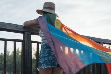 Unrecognizable young woman turning her back leaning on a wooden bridge with a rainbow flag knotted...