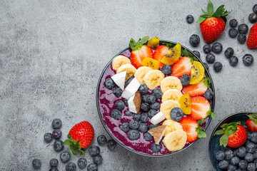 Healthy summer acai smoothie bowl with chia seeds, fresh banana, strawberry, blueberry, cocos, kiwi top view on rustic concrete background with spoon. Space for text