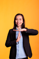 Cheerful positive model doing timeout symbol with hands, expressing half time and break in studio over yellow background. Filipino woman in formal suit requesting pause, gesturing rejection sign.