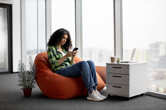 Pretty african american lady in everyday clothes texting messages on mobile while taking a seat on bean bag near file storage. Mindful freelancer writing emails via phone app in creative workplace.