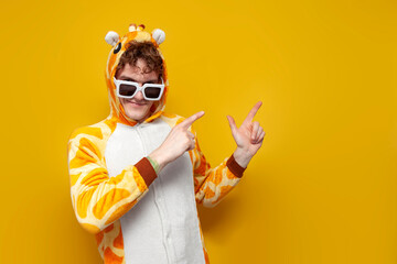 young joyful guy in funny baby giraffe pajamas and glasses points with his hands to the side on...