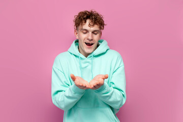 young curly guy in mint hoodie holds empty hands in front of him on pink isolated background, man...
