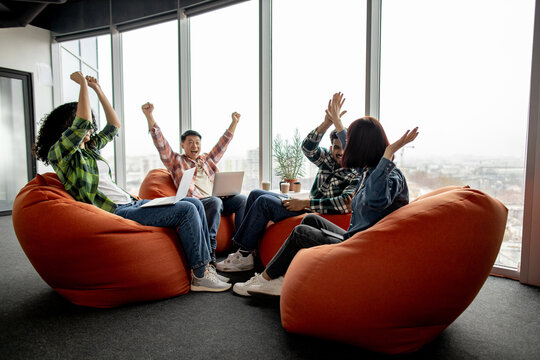 Diverse team of excited professionals in street style clothes raising fists in air and giving high five in workspace interior. Happy managers with devices celebrating successful project launch.