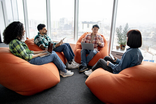 Diverse businesspeople holding mobile gadgets while taking seats on terracotta pouf chairs in airy coworking space. Young remote employees in street style outfits discussing corporate issues.