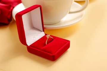 Box with engagement ring and cup of coffee on beige background, closeup
