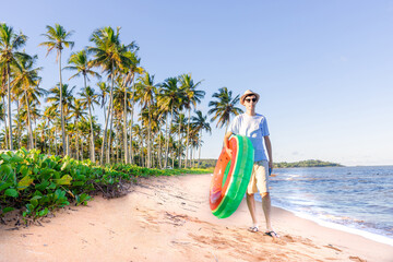  Man walking on the beach sand and carrying a watermelon float, with coconut trees in the background. Beach tourism.