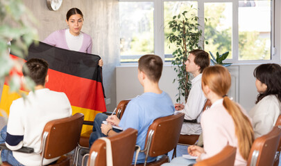 Positive young female student with interest telling group of coursemates about Germany at lecture, showing country national flag - obrazy, fototapety, plakaty