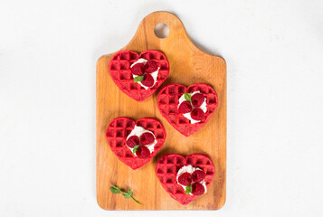 Small Red heart-shaped waffles with cream cheese, raspberries and micro greenery. Waffles with the addition of beet juice. On a serving wooden board. White background.  Top view