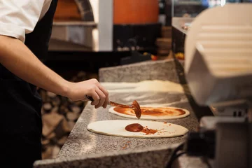 Foto op Plexiglas Anonymous male chef spreading tomato sauce with big iron spoon onto pizza while preparing pizza in pizzeria kitchen. High quality photo © arthurhidden