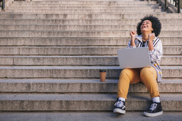 excited afro woman celebrating good news in laptop, sat in urban stairs