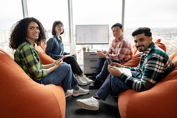 Four smiling business partners posing with handheld devices in coworking space with interactive...