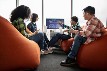 Diverse team of professionals utilizing smart gadgets while being busy at meeting in coworking space with soft poufs. Automotive engineers in casual wear analyzing digital data on display in office.