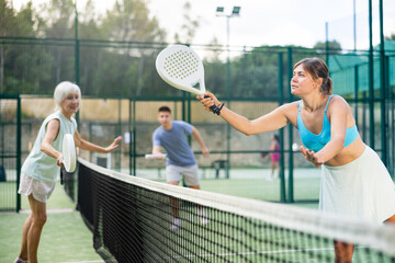 Young woman in skirt playing padel tennis on court with senior woman. Racket sport training outdoors.