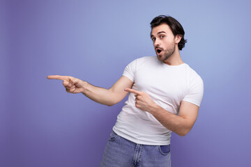 surprised brunette young man in a white t-shirt shows his index finger to the side on a studio background with copyspace